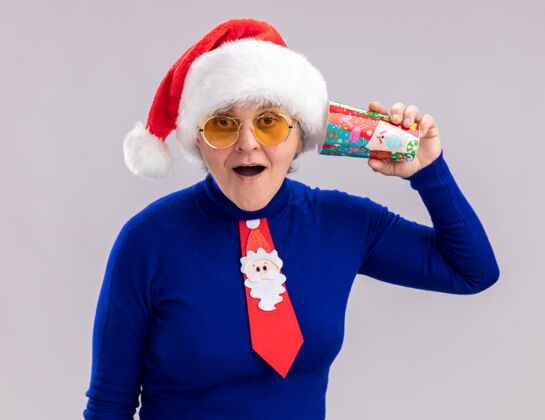 耳朵戴着太阳眼镜 戴着圣诞帽 系着圣诞领带的兴奋的老妇人把纸杯放在耳边兴奋纸女人