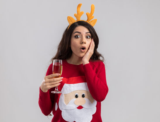 快乐惊讶的年轻漂亮女孩戴着驯鹿鹿角头带和圣诞老人毛衣拿着一杯香槟看着手放在脸上漂亮看圣诞老人
