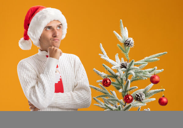 近戴着圣诞帽 系着圣诞老人领带的年轻帅哥抱着怀疑的态度站在装饰好的圣诞树旁 孤零零地仰望橘色的墙上圣诞老人圣诞老人年轻