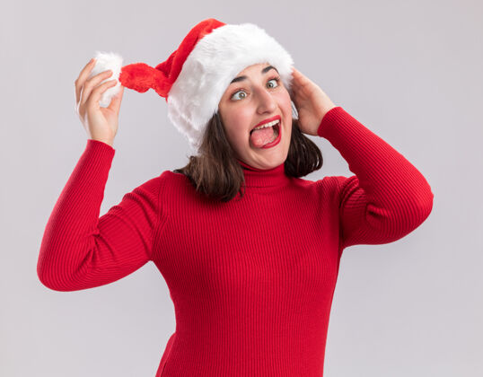 圣诞老人穿着红毛衣 戴着圣诞帽 站在白墙上做鬼脸伸出舌头的滑稽女孩站着毛衣做鬼脸