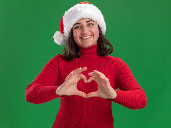 制作穿着红色毛衣 戴着圣诞帽的年轻女孩微笑着 手指站在绿色的墙上做着心形手势圣诞老人年轻人毛衣