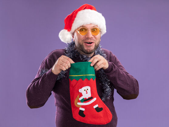 中年令人印象深刻的中年男子戴着圣诞帽 脖子上戴着金箔花环 戴着眼镜 手里拿着圣诞长袜 隔离在紫色的墙上帽子穿着男人