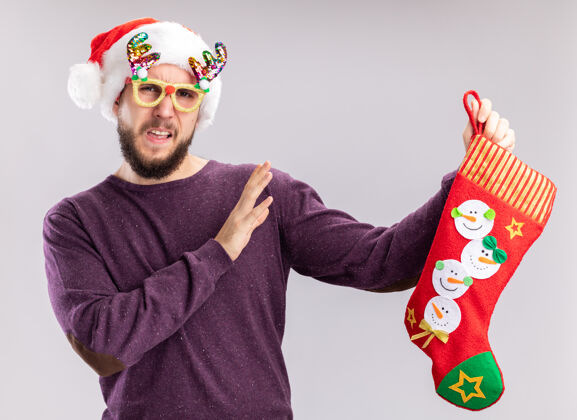 长袜一个穿着紫色毛衣 戴着圣诞帽 戴着滑稽眼镜 手里拿着圣诞长袜的年轻人困惑地站在白色的墙上做着防御的手势困惑年轻毛衣