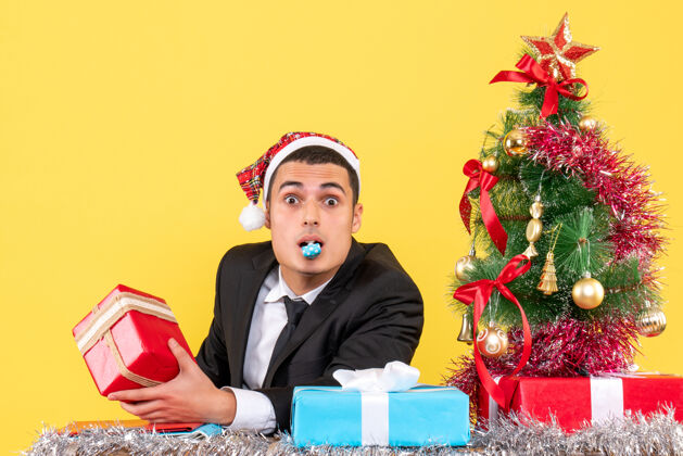成人前视图漫步的男子戴着圣诞帽坐在桌子旁拿着礼物用噪音制造圣诞树和礼物圣诞节男人礼物