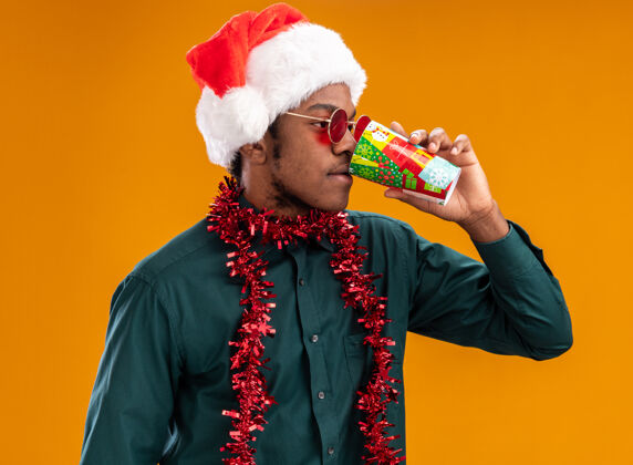 帽子戴着圣诞帽 戴着花环 戴着墨镜的非裔美国人站在橙色的墙上 喝着五颜六色的纸杯圣诞老人喝酒戴着