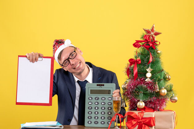 礼物商务人士与圣诞帽举行剪贴板和计算器坐在圣诞树附近的桌子上 黄色的礼物前视图剪贴板坐着乐器