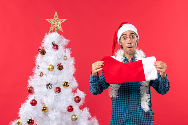 圣诞老人兴奋的年轻人 戴着圣诞老人帽 穿着蓝色的衬衫 手里拿着圣诞袜 站在圣诞树旁袜子庆祝礼物
