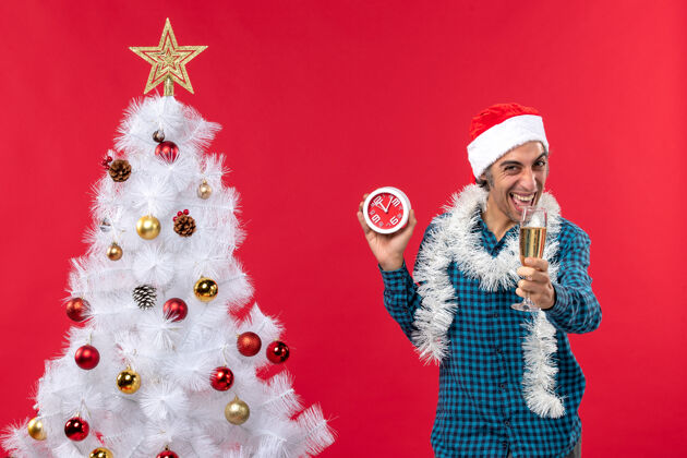 礼物笑容可掬的年轻人 戴着圣诞老人的帽子 举着一杯葡萄酒 拿着一个挂钟 站在红色的圣诞树旁圣诞老人抚养帽子