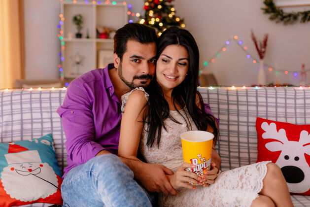 年轻一对年轻漂亮的情侣坐在沙发上 一男一女拿着一桶爆米花在装饰好的房间里一起看电视 背景是圣诞树一起电视爆米花