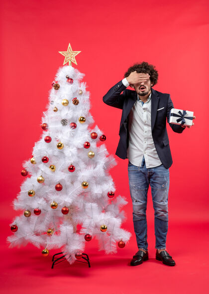 年轻人震惊的年轻人拿着他的礼物闭上眼睛站在红色的右侧装饰白色圣诞树附近树一边庆祝