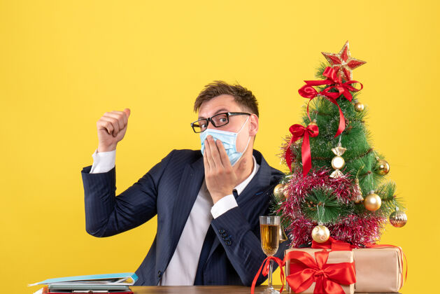 圣诞节前视图：商人把手放在嘴上 坐在圣诞树旁的桌子旁 黄色的礼物树职业黄色