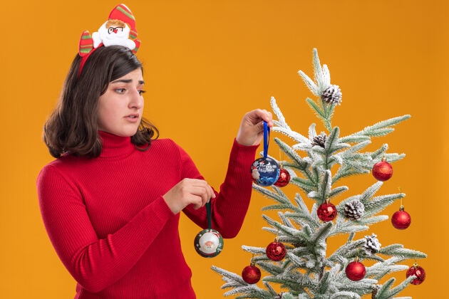 圣诞节困惑的小女孩穿着圣诞毛衣戴着有趣的头带旁边的圣诞树橙色背景戴着搞笑困惑