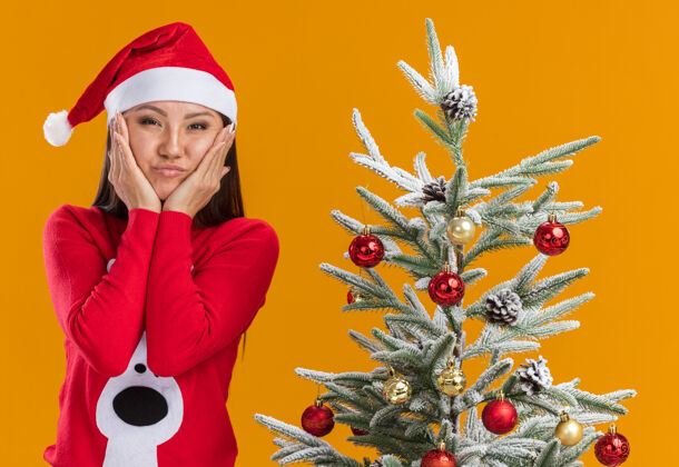 手高兴的年轻亚洲女孩戴着圣诞帽 穿着毛衣站在圣诞树旁 手放在橘色背景下孤立的脸颊上女孩穿着脸颊