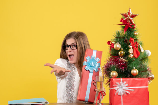 人正面图：女医生坐在桌子前 拿着礼物 黄色背景上的圣诞树和礼盒健康桌子女性