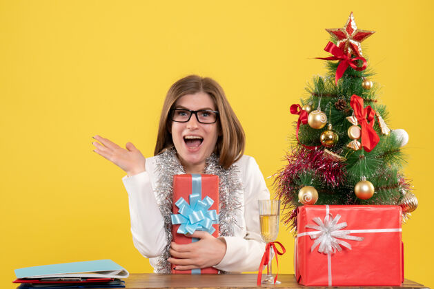 医生正面图：女医生坐在桌子前 拿着礼物 黄色背景上的圣诞树和礼盒坐着背景礼物