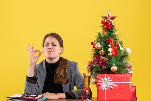 圣诞节正面图满意的女孩坐在桌子旁做圣诞树和礼物鸡尾酒旁的okey标志花束插花花