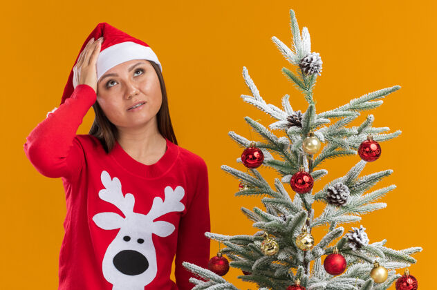 年轻人疲惫的年轻亚洲女孩戴着圣诞帽 穿着毛衣站在圣诞树旁 手放在孤立的橙色背景上的额头上手额头亚洲人