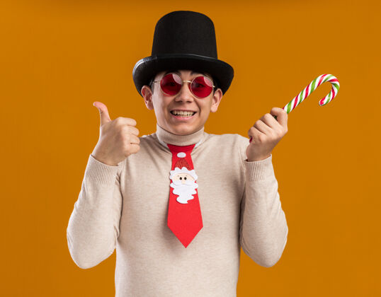 帽子微笑的年轻人戴着帽子 打着圣诞领带 戴着眼镜 手里拿着圣诞糖果 在黄色的墙上孤立地竖起大拇指糖果领带展示