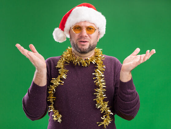 看笨手笨脚的中年男子戴着圣诞帽 脖子上戴着金箔花环 戴着眼镜 看着镜头 两只空手孤立在绿色背景上帽子男人相机
