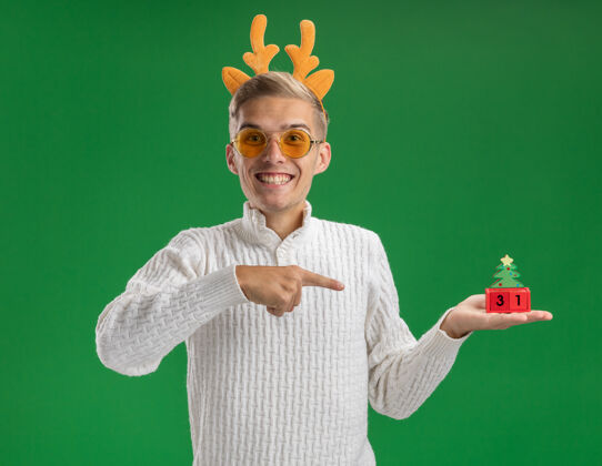 驯鹿快乐的年轻帅哥戴着驯鹿鹿角头带 戴着眼镜 拿着并指着绿色墙上的圣诞树玩具 上面的日期被隔离了快乐年轻指向