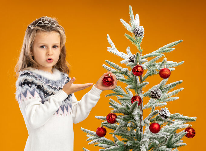 球困惑的小女孩站在圣诞树旁 戴着头饰 脖子上戴着花环 指着孤立在橙色背景上的圣诞球圣诞树圣诞头饰