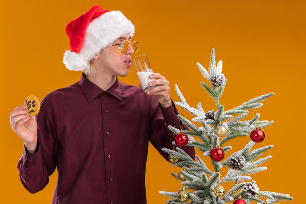 帽子戴着圣诞帽 戴着圣诞眼镜的年轻金发男子站在装饰好的圣诞树旁 嘴里端着一杯牛奶 眼睛紧闭的小甜饼被隔离在橙色的墙上金发饼干牛奶