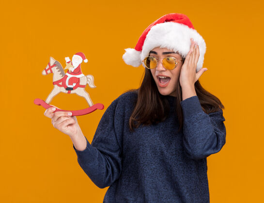 圣诞快乐惊讶的年轻白人女孩戴着太阳眼镜 戴着圣诞老人帽 手放在头上 手举着摇马 看着圣诞老人 橙色背景上的装饰与复制空间隔离开来头年圣诞节