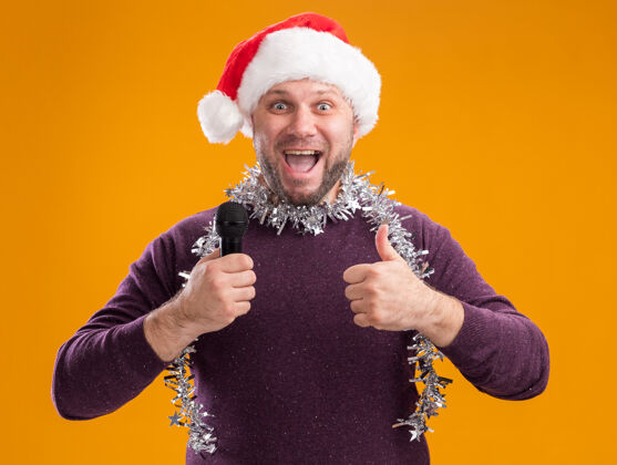 脖子令人印象深刻的中年男子戴着圣诞帽 脖子上戴着金箔花环 手持麦克风 在橙色的墙上孤立地竖起大拇指麦克风拇指印象深刻