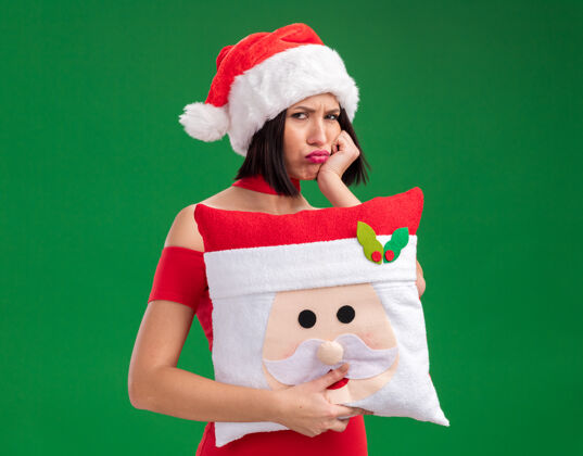 枕头闷闷不乐的年轻女孩戴着圣诞帽 抱着圣诞老人的枕头 手放在下巴上 隔离在绿色的墙上生闷气帽子下巴