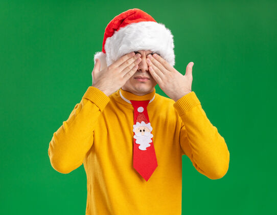 圣诞节戴着黄色高领毛衣和圣诞帽的年轻人 戴着滑稽的领带 双手站在绿色的墙上遮住眼睛领带年轻人男人