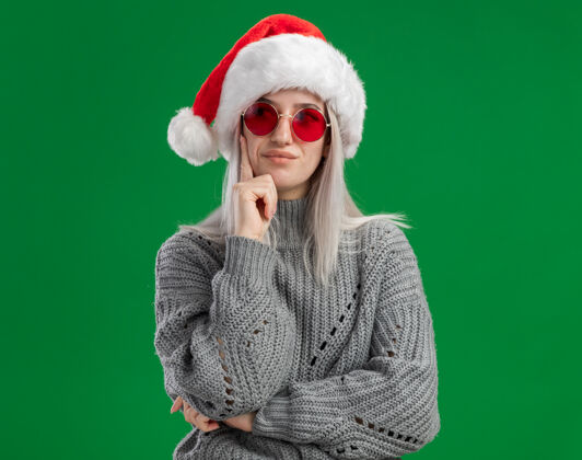 表情年轻的金发女郎 穿着冬衣 戴着圣诞帽 戴着红眼镜 站在绿色的背景下沉思着站着毛衣一旁