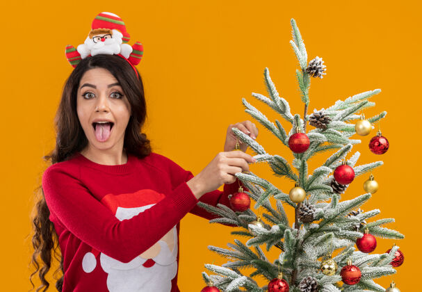 橙色顽皮的年轻漂亮女孩戴着圣诞老人的头带和毛衣站在圣诞树附近装饰圣诞树 看着相机显示舌头孤立在橙色的背景漂亮女孩站着
