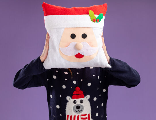 眼镜年轻漂亮的女孩穿着圣诞毛衣和帽子 戴着眼镜 脸上戴着圣诞枕头 背景是紫色的帽子脸毛衣