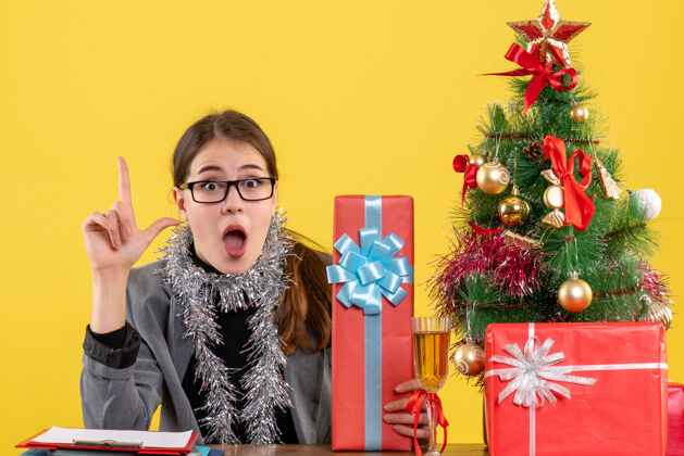 人前视图困惑的女孩戴着眼镜坐在桌边手指着圣诞树和礼物鸡尾酒树装饰女孩