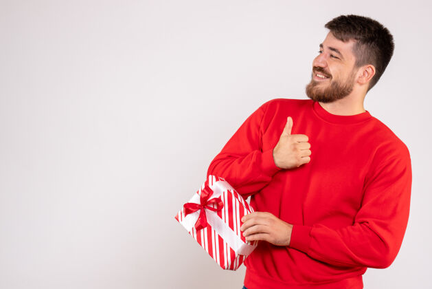 肖像身穿红衫的年轻人手持圣诞礼物站在白墙上的正视图前面微笑节日