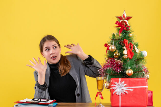 礼物前景色吓坏了坐在书桌旁张开双手的可爱女孩圣诞树和鸡尾酒礼物害怕人树