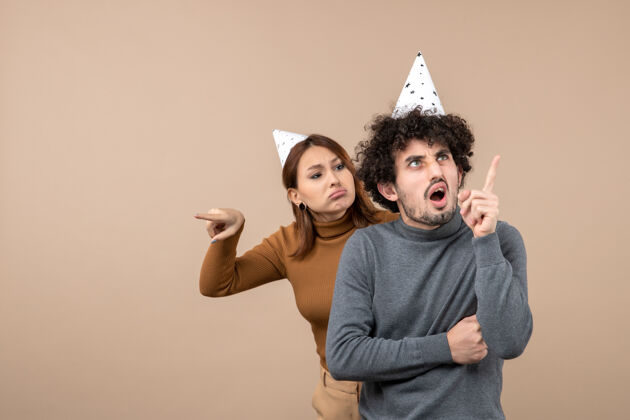 孩子可爱的年轻夫妇戴着新年帽愤怒的女孩看着站在后面的家伙指着灰色上面愤怒的年轻的夫妇指着