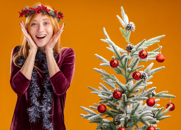 站着兴奋的年轻漂亮女孩站在圣诞树旁 穿着红色的裙子 脖子上戴着花环 手拉着隔离在橙色墙上的脸兴奋圣诞节年轻