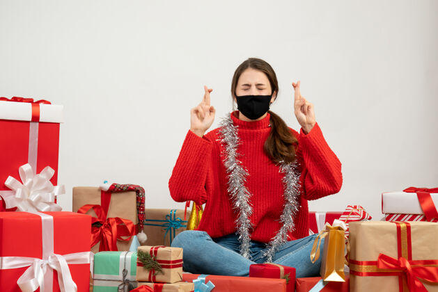 圣诞节穿着红色毛衣 戴着黑色面具的年轻女孩在白色的礼物上围坐着做好运的手势坐着毛衣黑色