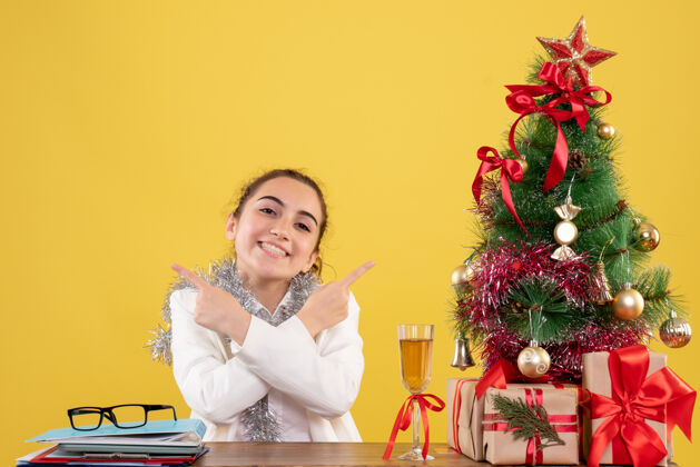 背景正面图：女医生坐在桌子后面 在黄色背景下微笑着 手里拿着圣诞树和礼品盒情感圣诞节成人
