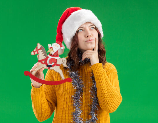 放体贴的年轻斯拉夫女孩戴着圣诞老人帽 脖子上戴着花环 把手指放在下巴上 把圣诞老人抱在摇椅上复制举行周围