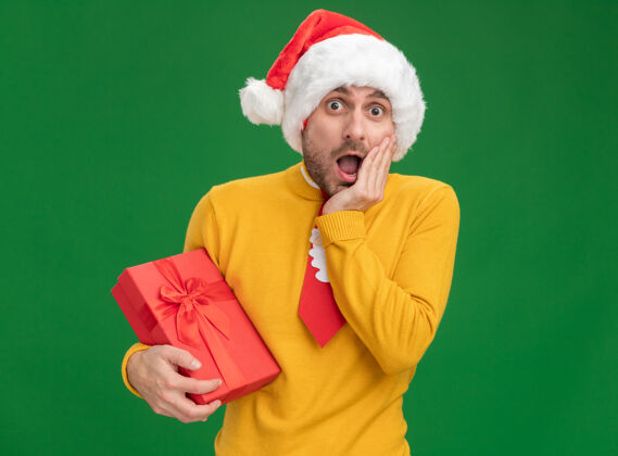 圣诞节关心的年轻白人男子戴着圣诞帽 打着领带 手里拿着礼包 手放在脸上 隔离在绿色的墙上 留着复印空间年轻关心男人