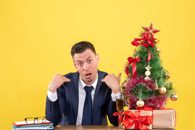 商人正面图是一个好奇的人指着自己坐在圣诞树旁的桌子 黄色的礼物指着桌子行政人员