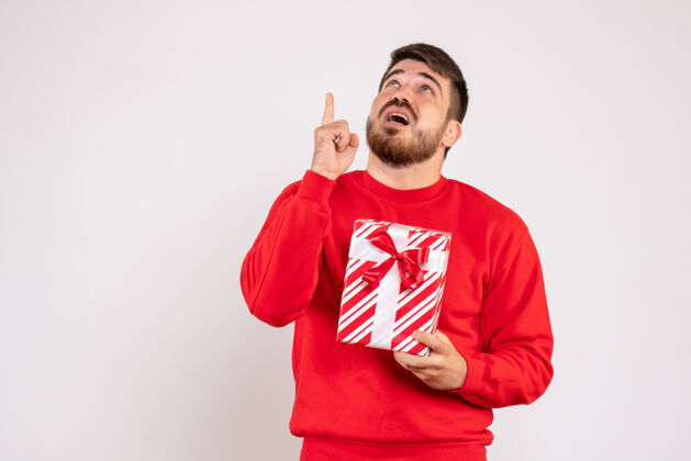 前面一个穿着红衫的年轻人在白色的墙上举着圣诞礼物的正视图情感现在男性