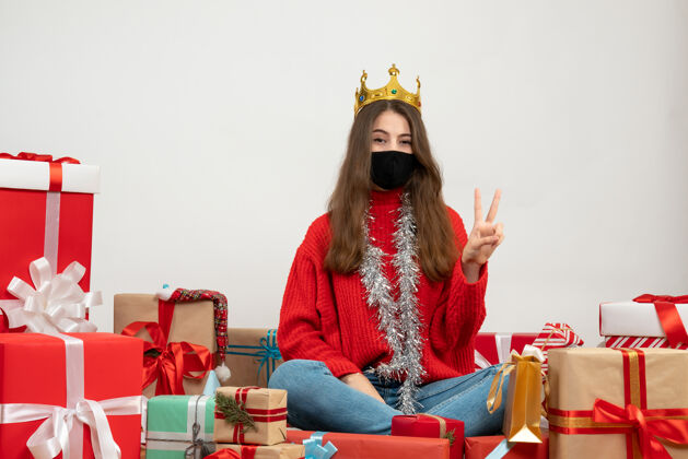礼物穿着红色毛衣的年轻女孩做着胜利的手势坐在礼物旁边 白色的面具上戴着黑色的面具周围红色坐着