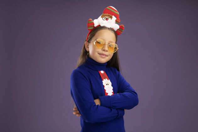 自信小女孩穿着蓝色高领毛衣 打着红色领带 头上戴着有趣的圣诞戒指 看着相机 自信地微笑着 双臂交叉着站在紫色的背景上领带搞笑微笑