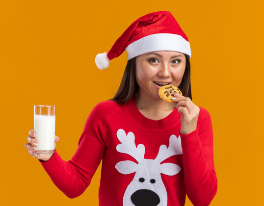 牛奶笑容满面的年轻亚洲女孩戴着圣诞帽 穿着毛衣 手里拿着一杯牛奶 在橙色的墙上品尝着孤立的饼干年轻人帽子毛衣