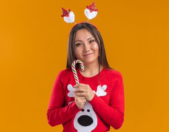 圣诞节高兴的年轻亚洲女孩戴着圣诞发箍和毛衣拿着圣诞糖果隔离在橙色的墙上高兴年轻糖果