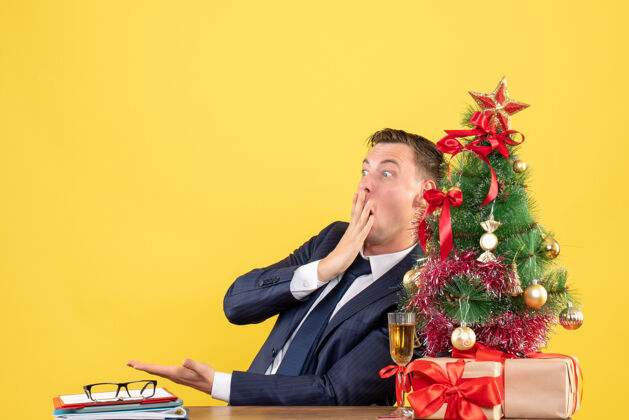 桌子前视图震惊的男子坐在圣诞树附近的桌子上 黄色的礼物笔记本电脑地方震惊的人