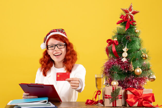圣诞节正面图：女医生坐在桌子后面 拿着黄色背景的银行卡 还有圣诞树和礼品盒情绪抱着背景